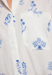 Biała koszula z niebieskim kwiatowym haftem, z kieszonką i zapięciem na guziki, wykonana z miękkiego materiału o gładkiej teksturze.