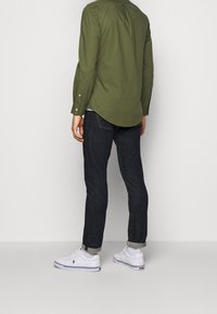 Mężczyzna w podwiniętych ciemnych dżinsach, białych trampkach, białych skarpetkach i długim rękawie oliwkowej zielonej koszuli stojący na tle gładkiej jasnej ściany.