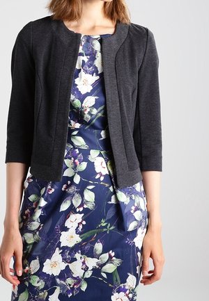 Vrouw draagt een donkergrijs cropped vest over een marineblauwe jurk met een wit-groene bloemenprint, staand tegen een effen lichte achtergrond.