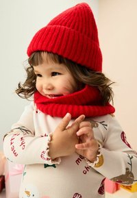 Un jeune enfant porte un bonnet en tricot rouge et une écharpe assortie, souriant avec les mains jointes. L'arrière-plan présente des couleurs douces et des boîtes cadeaux.