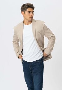 Beige blazer med en enkelt brystlomme, parret med en hvid T-shirt og mørkeblå jeans. Glat stof, struktureret design og skræddersyet pasform.