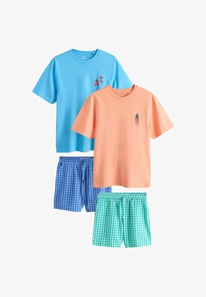 Mladý muž v svetlomodrej košeli s malým grafickým motívom paradajkovej rastliny na hrudi, stojaci pred jednoduchou svetlou stenou.