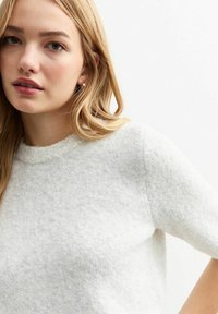 Vit kortärmad tröja med en mjuk, texturerad yta och rund halsringning, bärs av en person med långt hår och minimala accessoarer.