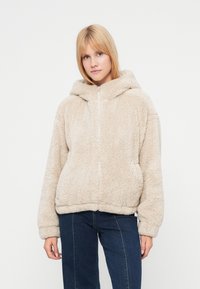 Μπεζ φλις φούτερ με φερμουάρ μπροστά, χαλαρή εφαρμογή και κουκούλα με κούμπωμα. Διαθέτει ριμπ μανσέτες και τελείωμα, μαλακή υφή και χωρίς ορατά σχέδια.