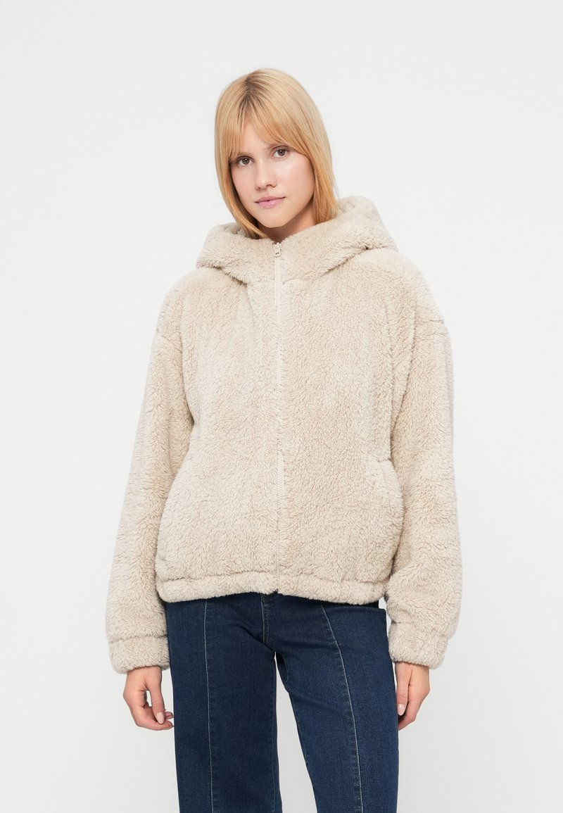 Μπεζ φλις φούτερ με φερμουάρ μπροστά, χαλαρή εφαρμογή και κουκούλα με κούμπωμα. Διαθέτει ριμπ μανσέτες και τελείωμα, μαλακή υφή και χωρίς ορατά σχέδια.