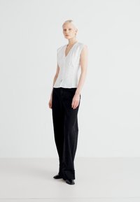 Blouse blanche sans manches avec un col en V et un ruché à l'avant, associée à un pantalon noir taille haute à jambes larges en tissu texturé.