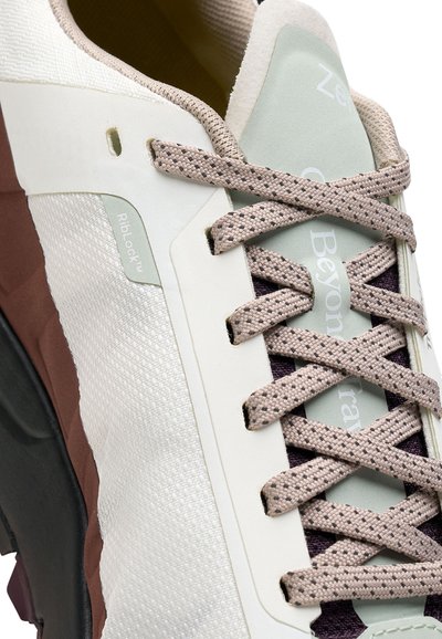 Gros plan sur des baskets beige et blanches avec des lacets à motifs, des côtés en tissu texturé et une semelle accentuée en marron. Le texte "RibLock" est visible sur le panneau latéral.