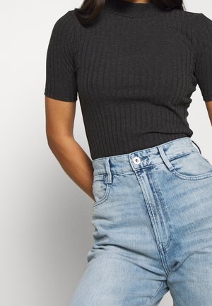 Woman wearing a fitted black ribbed short-sleeve top tucked into high-waisted light blue denim jeans with hands in pockets.