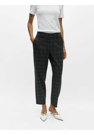Femme portant un pantalon court à carreaux gris foncé, un haut blanc à manches courtes et des chaussures blanches à bout pointu et talons bas avec des brides décoratives.