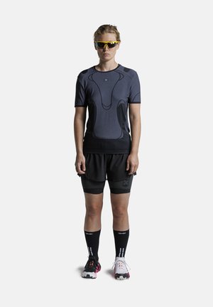 Femme portant un t-shirt de sport noir et gris, un short noir, des chaussures de course dépareillées, des chaussettes noires hautes et des lunettes de soleil de sport jaunes, debout immobile.