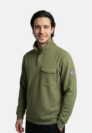 Mann mit dunklen Haaren, der einen olivgrünen Pullover mit einer geknöpften Brusttasche und einem kleinen Aufnäher am linken Ärmel trägt, steht und lächelt.