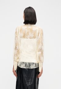 Μπλούζα από δαντέλα σε κρεμ χρώμα, με μακριά φουσκωτά μανίκια, λουλουδάτο σχέδιο και απλή λευκή πλάτη. Συνδυάζεται με μαύρη δερμάτινη φούστα.