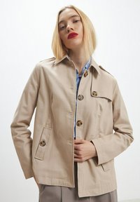 Giacca di cotone beige con chiusura a bottone, due tasche frontali e colletto classico. Presenta una texture liscia e una vestibilità su misura.