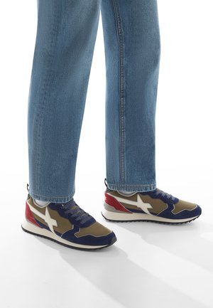 Persona che indossa jeans blu e sneakers multicolori con accenti blu navy, oliva, bianchi e rossi, in piedi su un pavimento bianco.