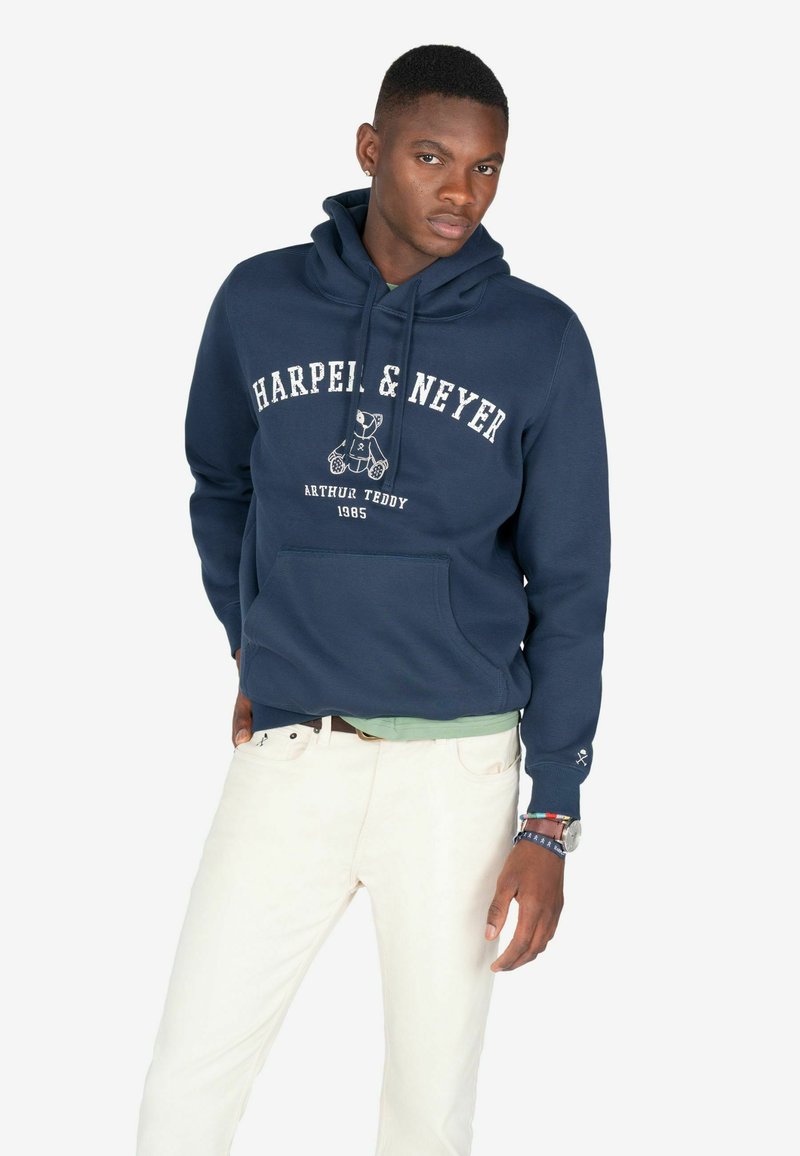 Granatowy hoodie z kieszenią z przodu, z białym napisem i grafiką misia. Połączony z jasnymi spodniami, prezentując swobodny wygląd.