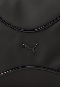 Svart väska med böjda linjer, tillverkad av mjukt material. Försedd med en glansig svart Puma-logotyp. Enkel och modern design.