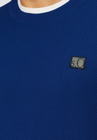 Blauer strukturierter Pullover mit Rundhalsausschnitt und einem kleinen schwarzen Logopatch auf der linken Seite. Das Strickmuster ist dezent und detailliert.