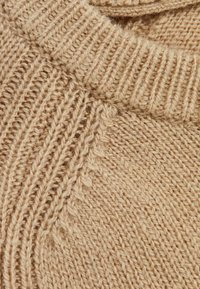 Prim-plan al unui țesut de lână bej tricotat, evidențiind gulerul cu nervuri și modelele texturate detaliate ale cusăturilor.