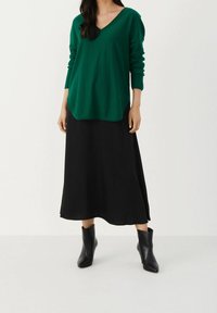 Vihreä V-pääntie neule, väljä malli pitkillä hihoilla, yhdistetty mustan maksihameen kanssa. Mustat nilkkurit. Sileät materiaalit, ei näkyviä kuvioita.