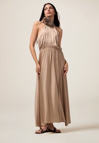 Pitkä, beige satiininen halter-mekko, jossa on röyhelökaulus ja joustava vyötärö, jota täydentävät naruilla kiinnitettävät mustat sandaalit.