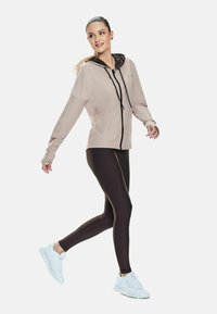 Young woman in beige zip-up hoodie, black leggings, and white sneakers walking with a smile on a plain white background.