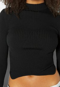 Černý žebrovaný pletený top s vysokým límečkem, přiléhavým střihem a trojúhelníkovým texturovaným detailem přes hruď. Krátká délka s hladkým lemem.
