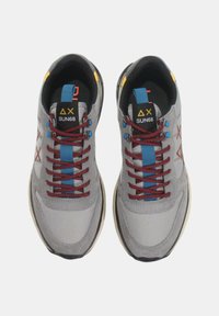 Sneaker grigie con tomaia in tessuto, lacci rossi e blu, dettagli gialli e suola bianca a contrasto. Logo distintivo e dettagli decorativi sui lati.