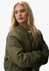 Oliwkowa kurtka bomber z zamkiem z przodu, z prążkowanymi mankietami i dołem, z naszywką z amerykańską flagą na lewym rękawie.