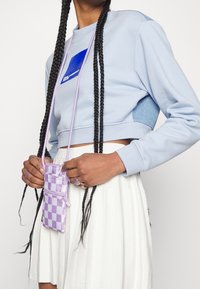 Jasnoniebieski skrócony sweatshirt z długimi rękawami, z niebieskim logo. Lawendowa mini torebka w kratkę noszona na ramieniu.