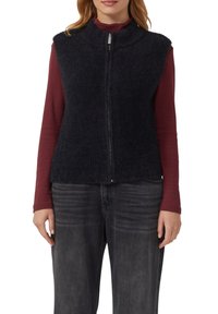 Marinefarbene, strukturierte Weste mit hochgeschlossenem Kragen und Vorderreißverschluss, übereinander getragen über einem roten Langarmoberteil. Kombiniert mit grauen Jeans.