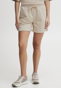 Shorts en coton beige avec une taille à cordon, des poches latérales et une texture douce, associés à des baskets gris clair.