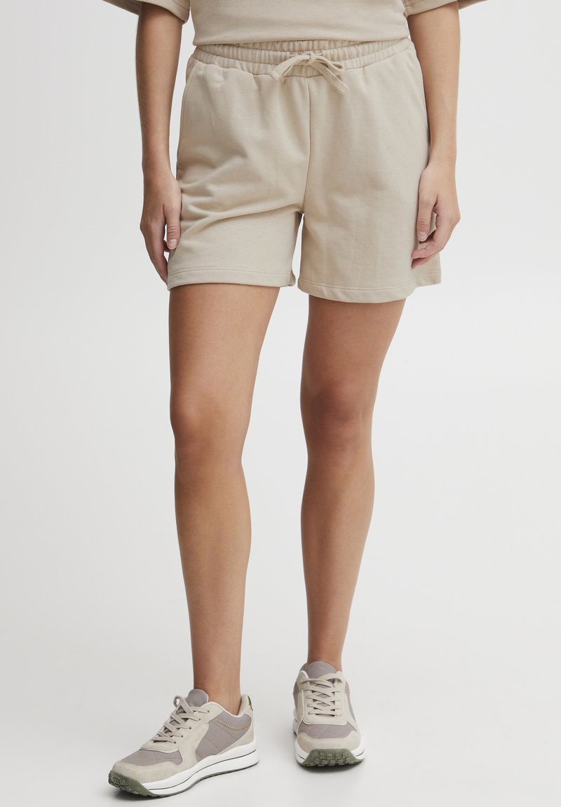 Shorts en coton beige avec une taille à cordon, des poches latérales et une texture douce, associés à des baskets gris clair.