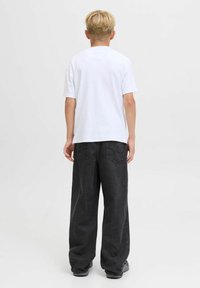 Une personne aux cheveux blonds portant un t-shirt blanc simple, un jean noir ample et des baskets grises, se tenant face à un fond blanc.