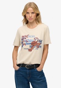 Beżowa bawełniana koszulka z kolorowym nadrukiem, przedstawiającym motywy kwiatowe i tygrysy, okrągłym dekoltem i krótkimi rękawami, w połączeniu z niebieskimi dżinsami.