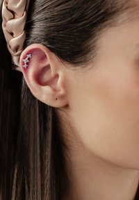 Srebrny klips na ucho z małymi, przezroczystymi kryształkami ułożonymi wzdłuż zewnętrznej krawędzi ucha. Widoczna gładka karnacja oraz ciemne, proste włosy.