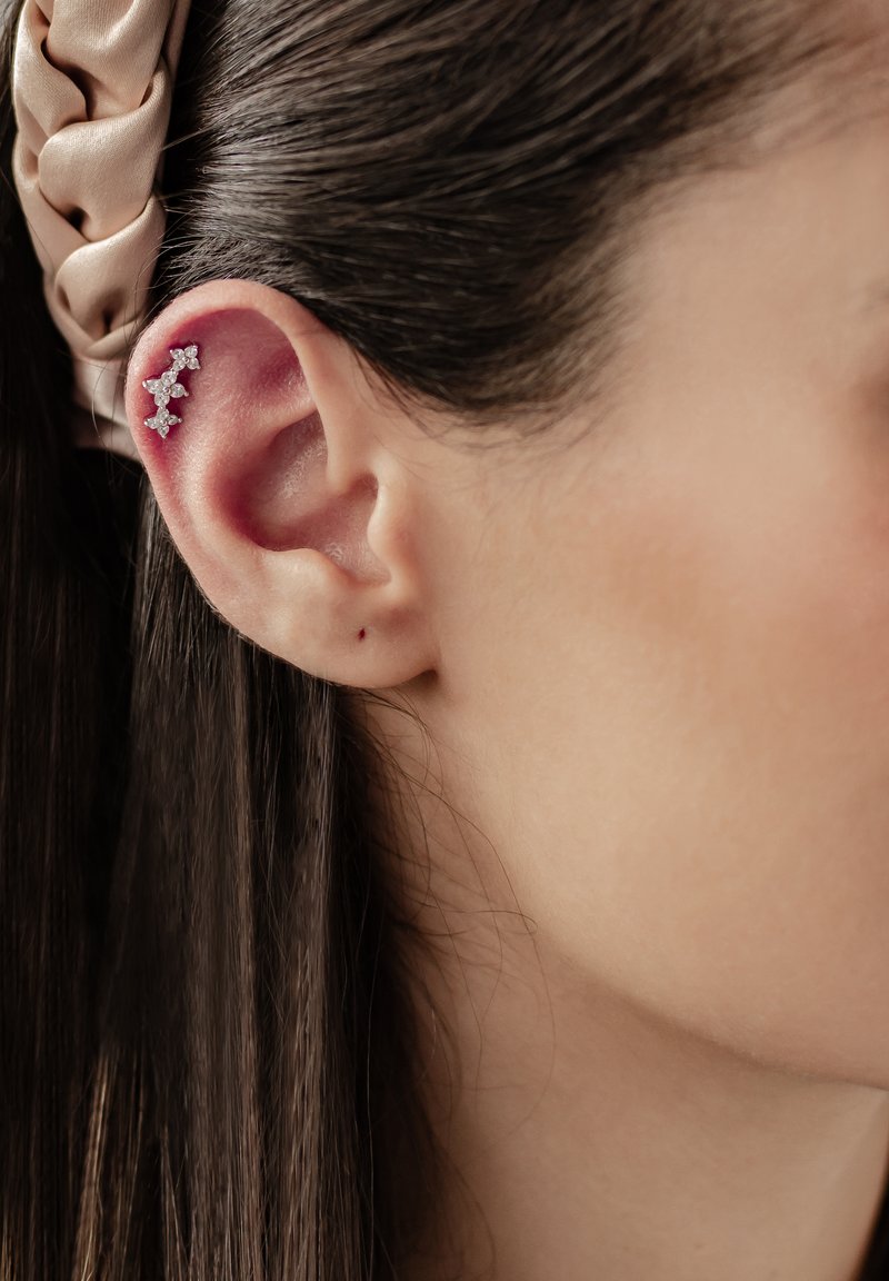 Srebrny klips na ucho z małymi, przezroczystymi kryształkami ułożonymi wzdłuż zewnętrznej krawędzi ucha. Widoczna gładka karnacja oraz ciemne, proste włosy.