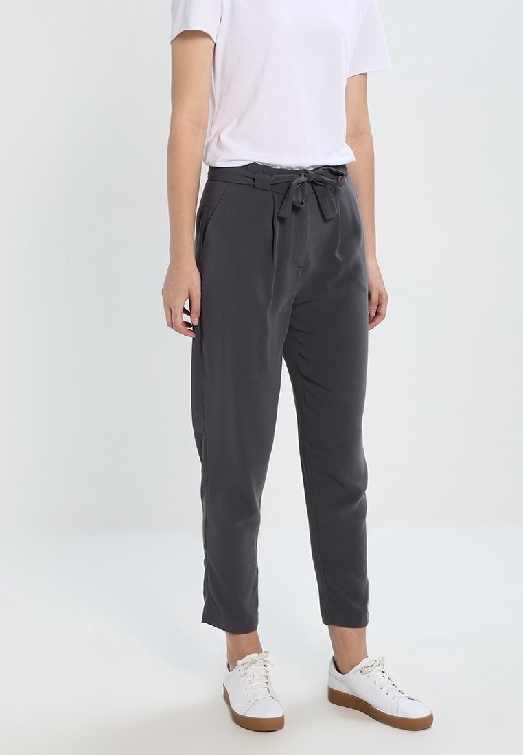 Personne portant un pantalon taille haute gris foncé à effet sac avec ceinture nouée, un t-shirt blanc et des baskets blanches à semelles marron, debout sur un sol blanc.