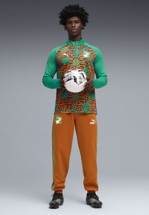 Hombre con chaqueta deportiva de patrón verde y naranja y pantalones naranja sostiene un balón de fútbol en blanco y negro, de pie contra un fondo gris.