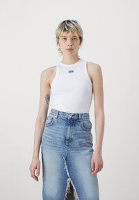 Vit ärmlös linne med rund halsringning och blå logotyp, kombinerad med en ljusblå denimkjol med slits fram och fransad nederkant.