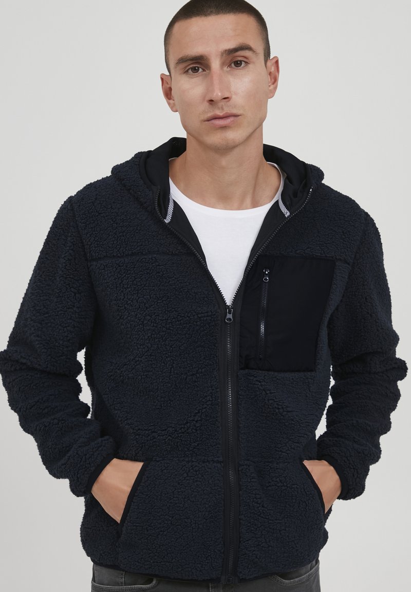 Navy blue fleece hoodie with a textured finish, contrasting black zippered chest pocket, full zipper, and side hand pockets.