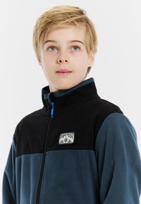 Fleece jack in blauw en zwart, met een hoge kraag, ritssluiting en een logo patch op de borst. Zachte textuur, casual ontwerp.