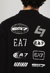Black jacket featuring various white EA7 logos and a globe design on the back. The fabric has a textured appearance with a modern cut.