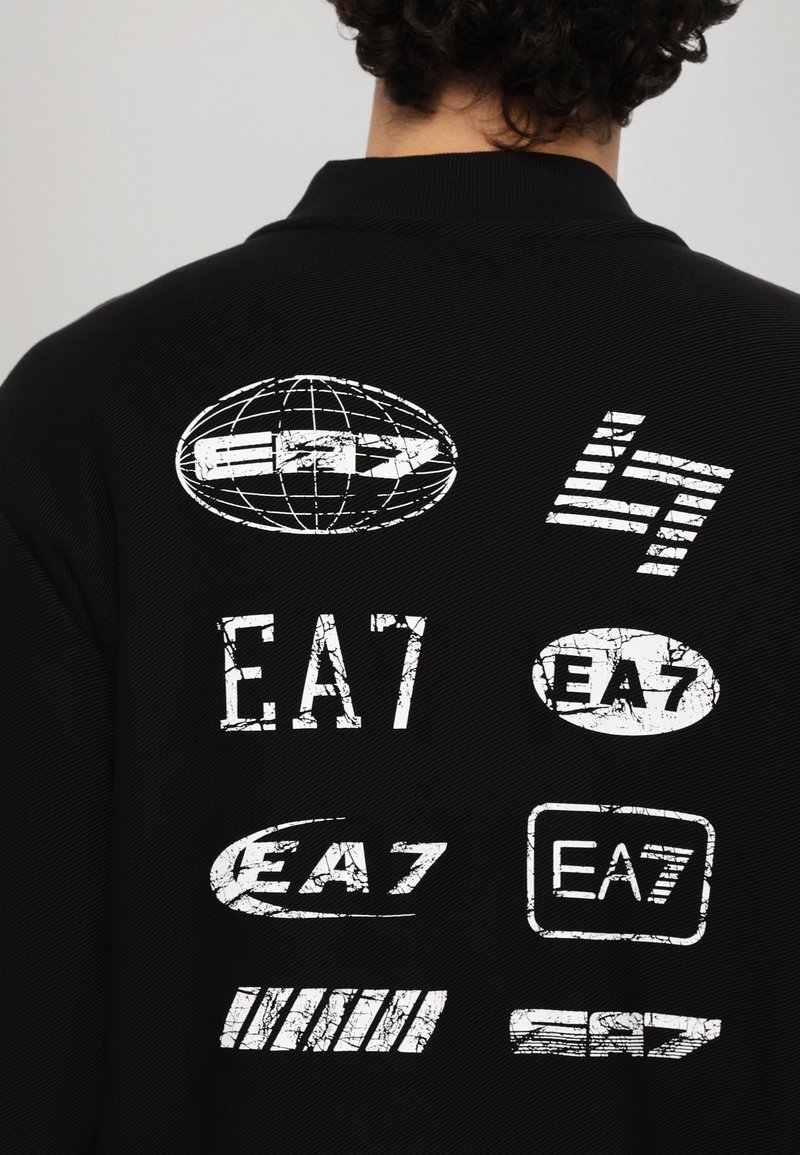 Chaqueta negra con varios logotipos blancos de EA7 y un diseño de globo en la parte posterior. La tela tiene una apariencia texturizada con un corte moderno.