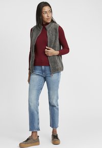Graue Fleece-Weste mit hohem Kragen, über einem burgunderfarbenen Rippensweater getragen, kombiniert mit hellblauen geraden Jeans und gemusterten Schuhen.