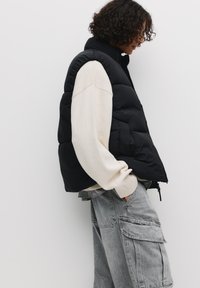 Gilet trapuntato nero con colletto alto, indossato sopra un maglione color crema. Presenta una chiusura con zip e tasche laterali, abbinato a pantaloni cargo grigi.