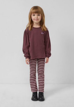 Een jong meisje staat rechtop en draagt een wijnrood shirt met lange mouwen met een klein bloemenpatroon, gestreepte broek en zwarte laarzen, tegen een effen achtergrond.