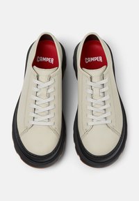 Sneaker beige con suole nere e lacci bianchi, con solette rosse etichettate "CAMPER" su sfondo bianco semplice.