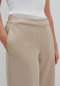 Beige elastische Hosen mit glatter Textur, ausgestattet mit Seitentaschen und einem lockeren Schnitt. Das Oberteil ist ein tailliertes weißes Ripp-Top.