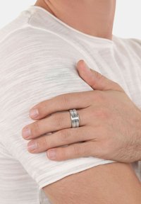 Anello argentato con tre fasce sovrapposte, caratterizzato da superfici testurizzate e finiture alternate tra lucide e opache. Indossato su una mano.