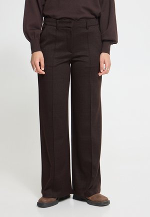 Femme portant un pantalon large taille haute marron foncé avec plis sur le devant et des chaussures marron, debout devant un fond clair uni.